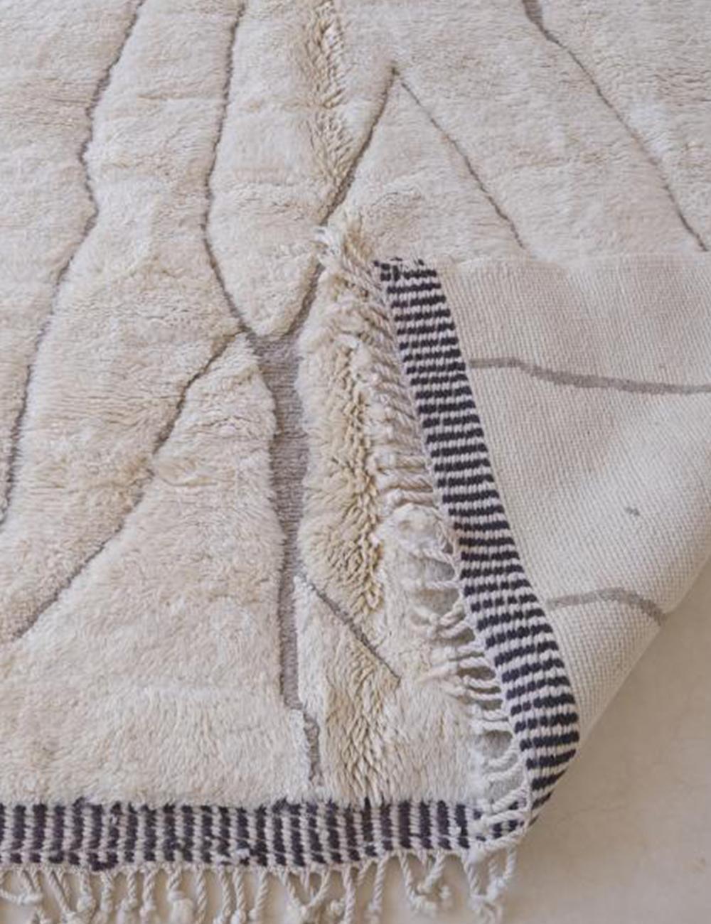 Contemporary Moroccan Beni Mrirt wool rug in beige with thin gray abstract lines, folded corner showing woven underside and fringed black-and-white border.