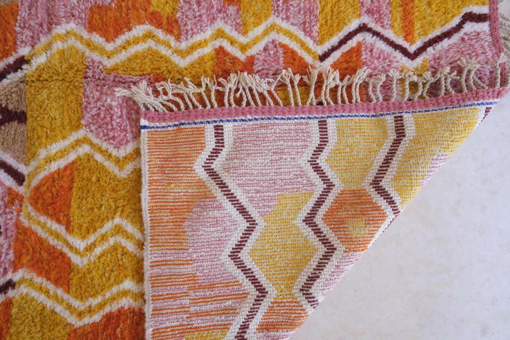 Modern Moroccan Boujad wool rug with colorful zigzag patterns in yellow, orange, and pink, folded corner showing woven underside and fringed edge.