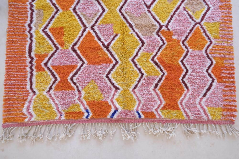 Detail of modern Moroccan Boujad wool rug with vibrant yellow, orange, and pink zigzag patterns and white fringed edge.