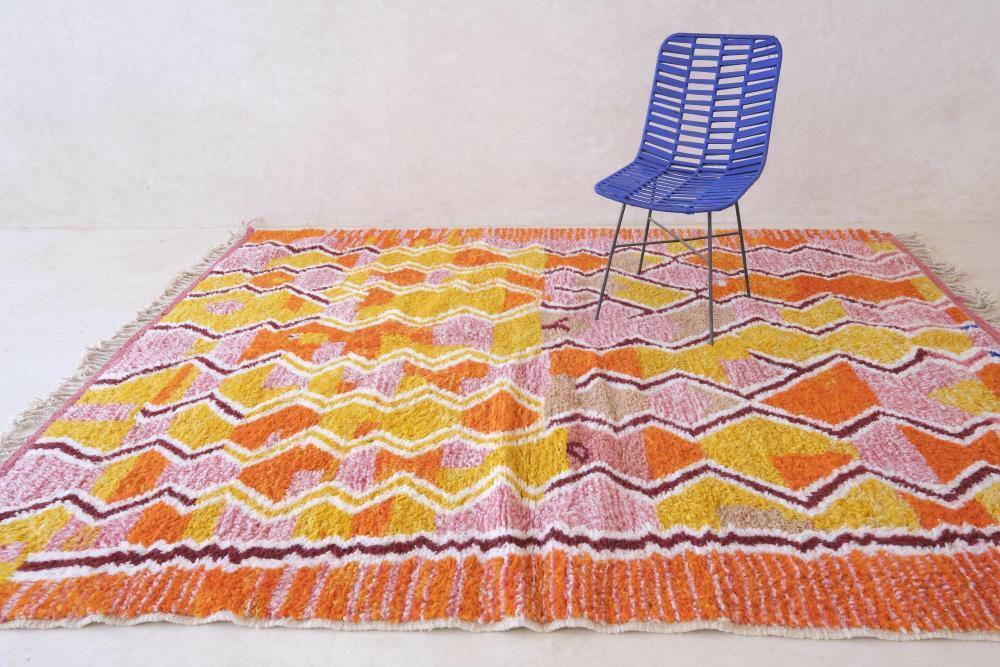 Modern Moroccan Boujad wool rug with vibrant pink, yellow, and orange zigzag patterns and fringed edges, styled with a blue chair.
