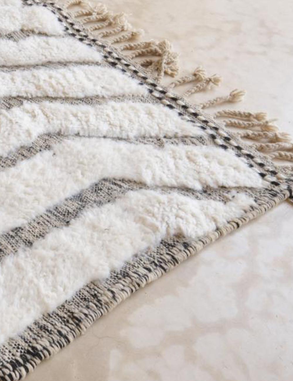 Corner of a Moroccan Beni Ourain wool rug with black geometric lines and tasseled fringe, placed on a light marble floor.