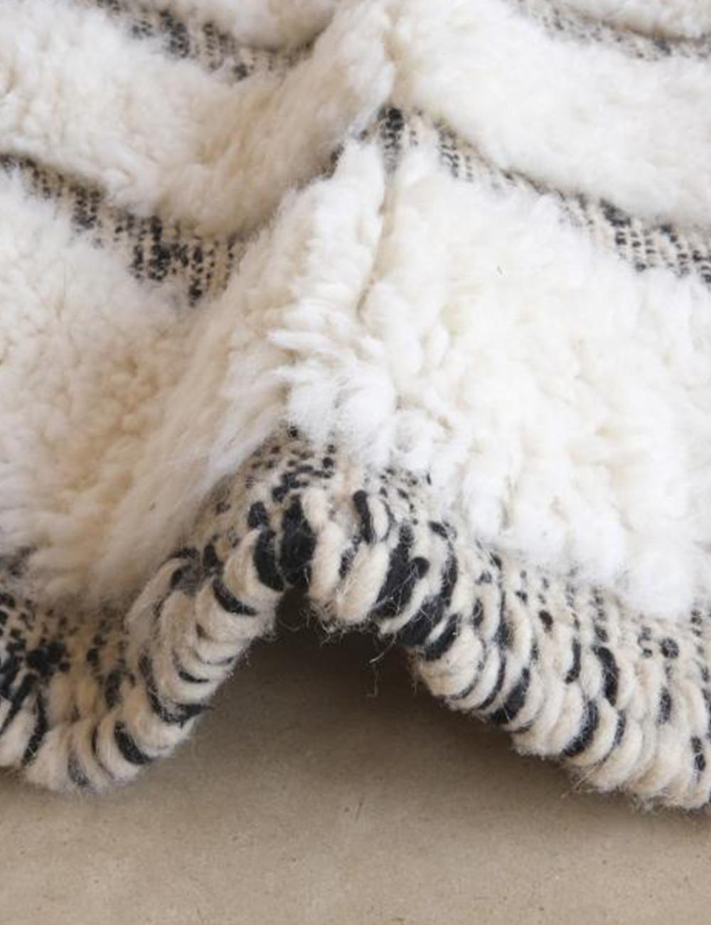 Close-up of a Moroccan Beni Ourain wool rug with thick white pile, black geometric pattern, and braided edge.