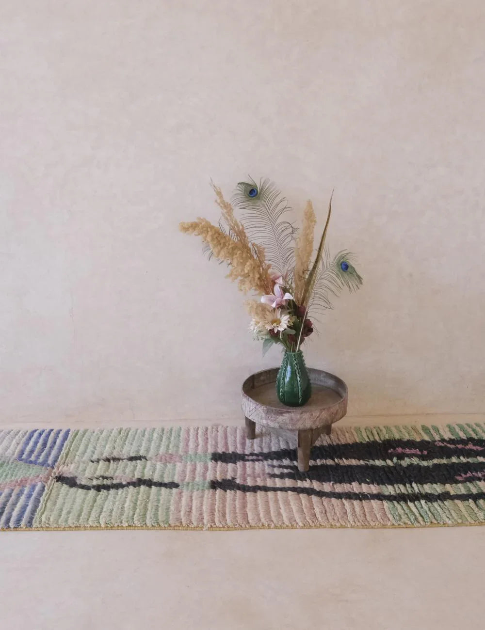 Small round wooden table with a vase of flowers on a colorful rug against a plain wall.