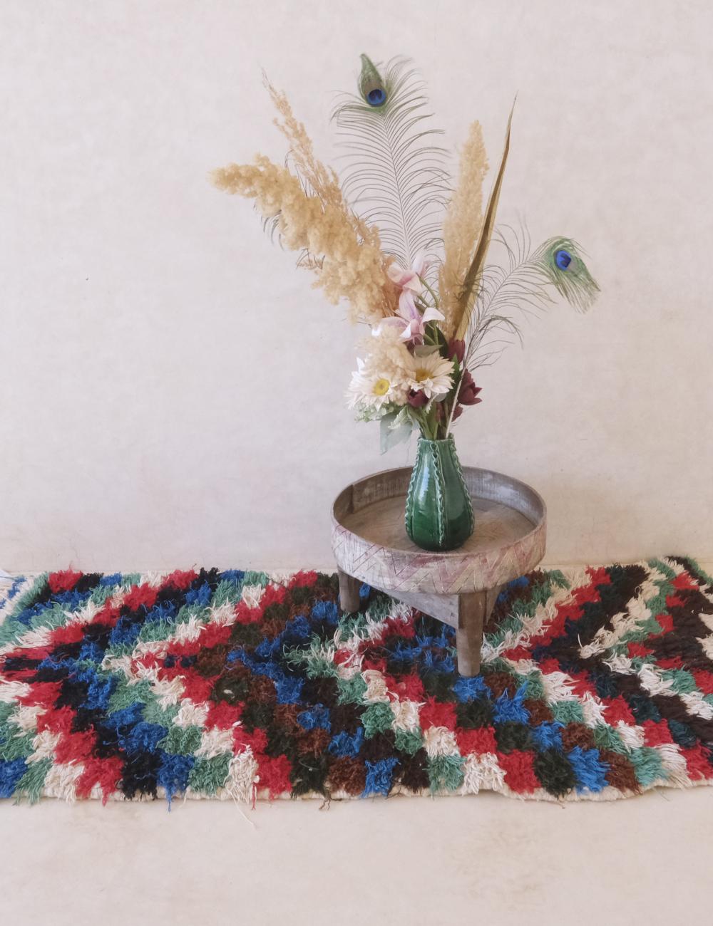 Colorful woven rug with a small round table holding a vase with flowers and peacock feathers.