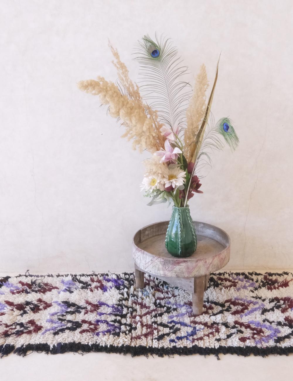 Vase with flowers and peacock feathers on a small wooden table against a white wall.