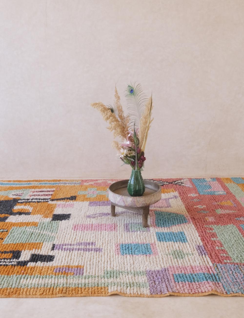 Colorful woven rug with a vase of flowers on a small table against a plain wall.