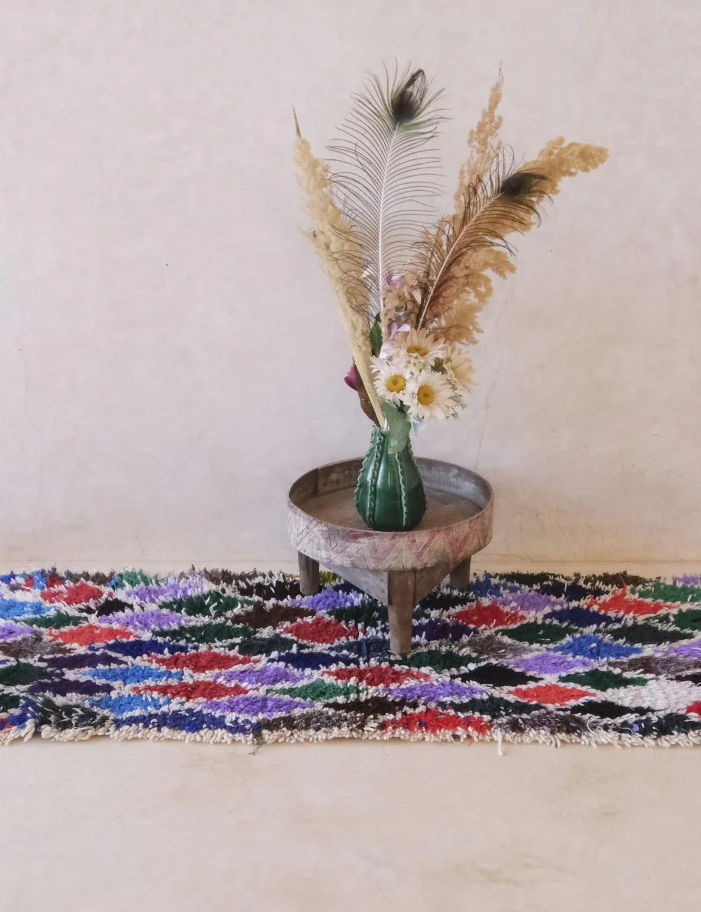 Decorative rug with a vase of flowers and feathers on a small table against a plain wall.