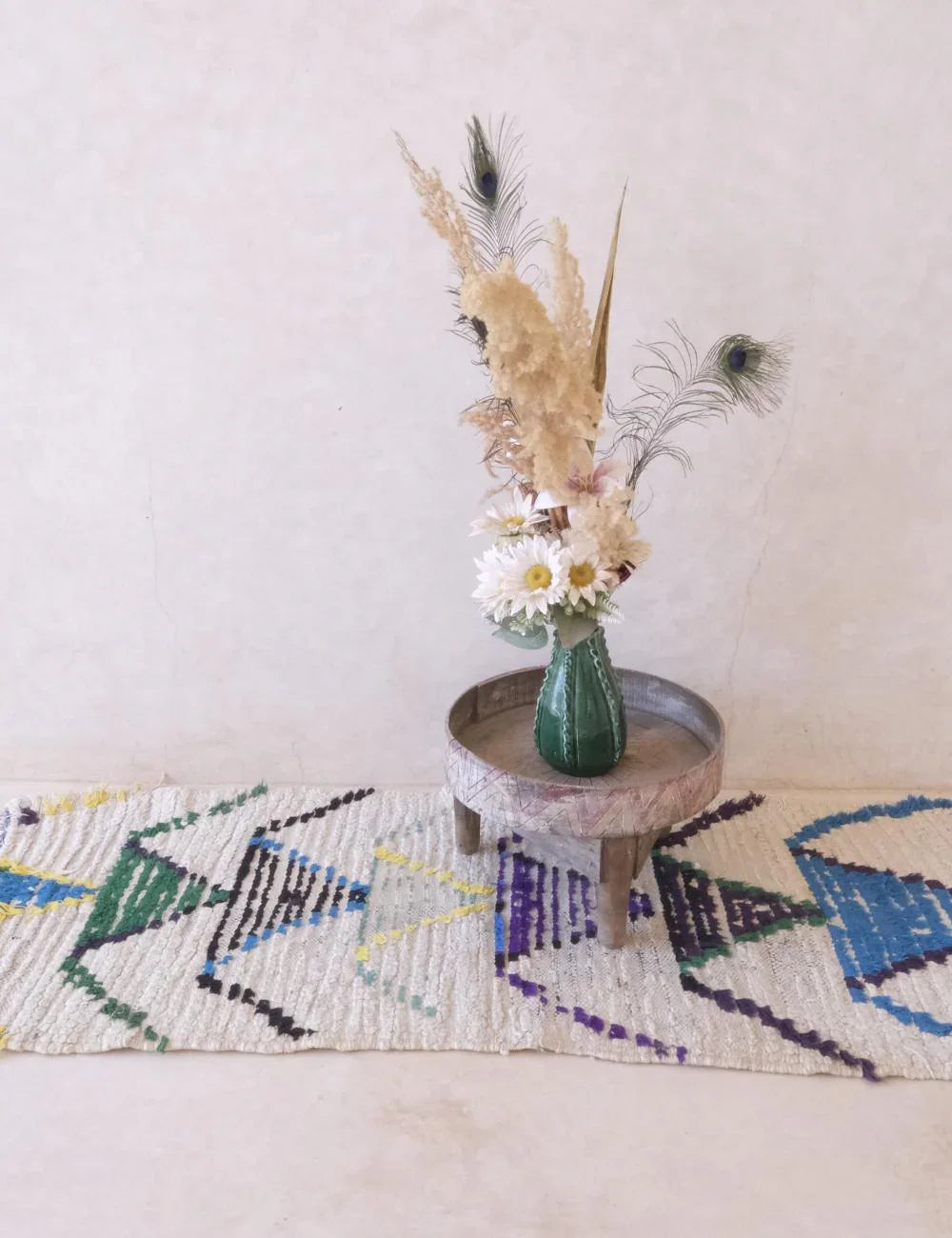 Decorative vase with flowers on a small round table against a light wall, with a colorful rug below.