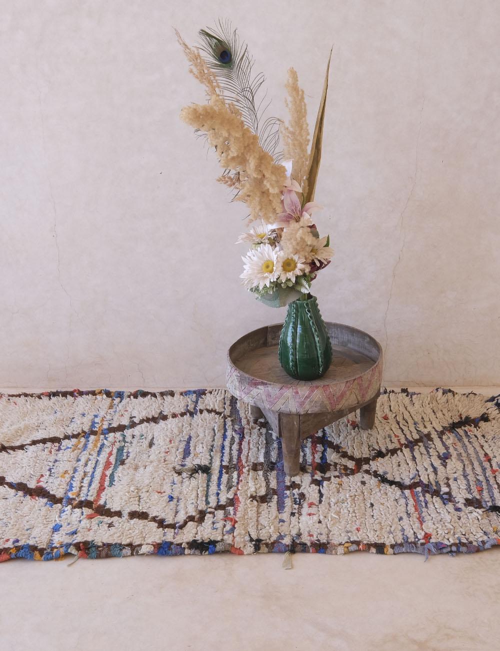 Decorative vase with flowers and peacock feathers on a small round table, with a colorful rug in the foreground.