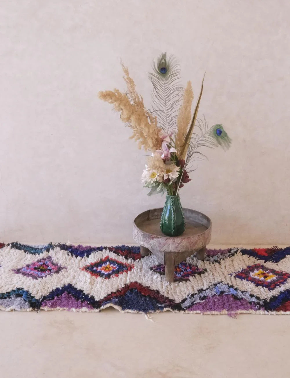 Decorative rug with a vase of flowers and peacock feathers on a wooden stool against a plain wall.