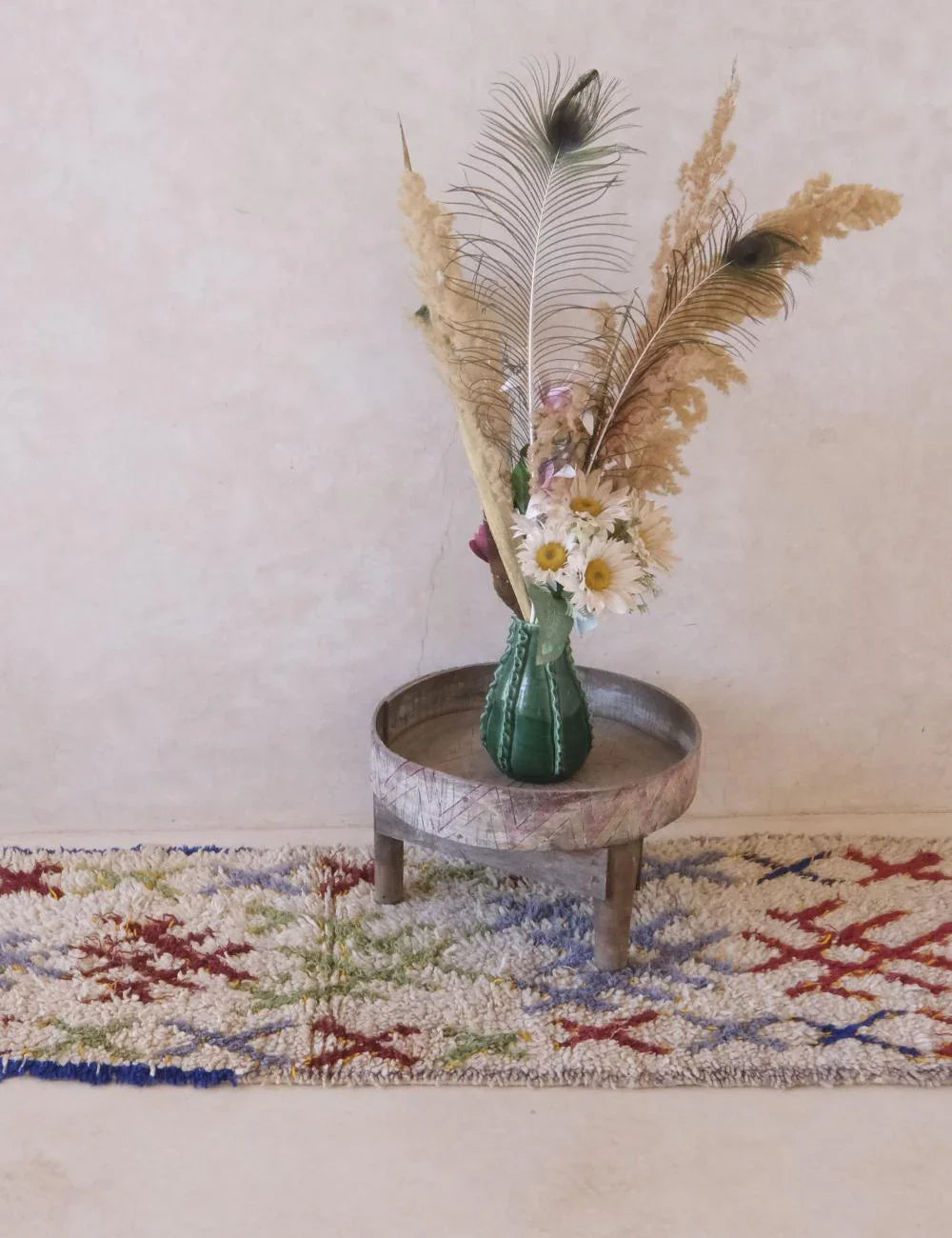 Decorative vase with flowers and feathers on a small round table against a plain wall.