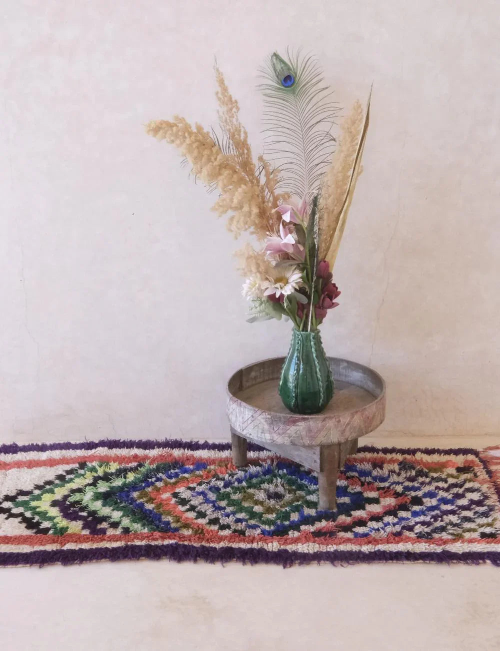 Decorative vase with flowers and peacock feather on a small round table with a colorful rug underneath.