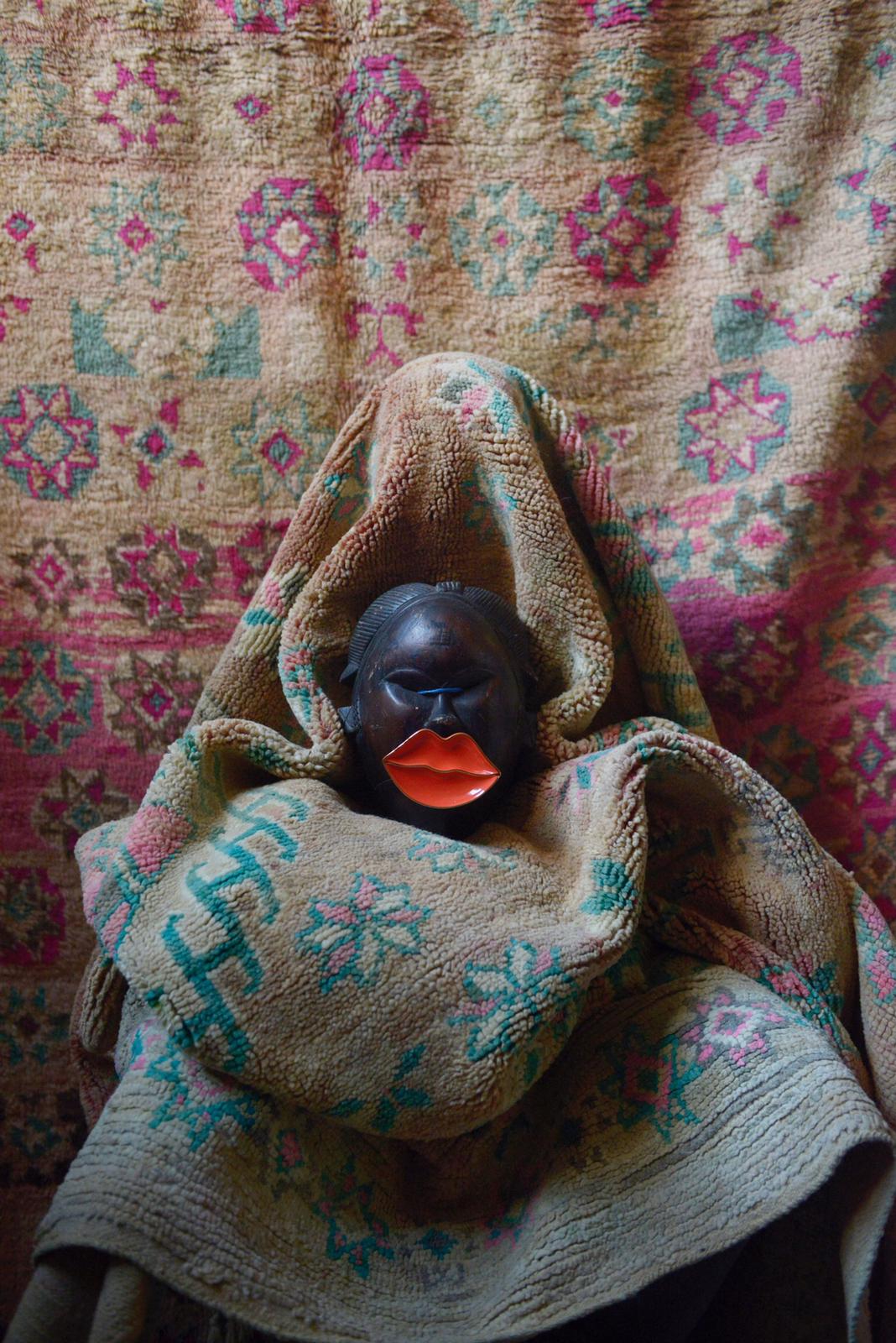Carved mask with white hair tufts draped in vintage Moroccan textile in front of colorful rug.