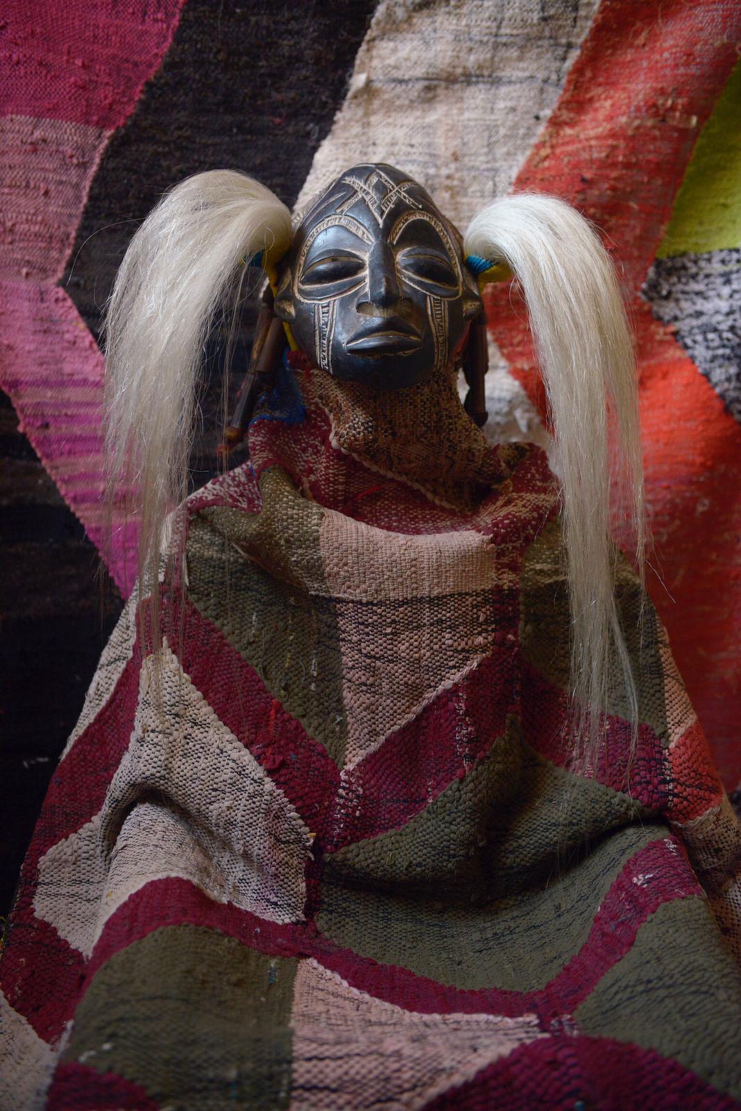 Wooden mask with red lips nestled in beige Boujad-style rug against floral pink backdrop.