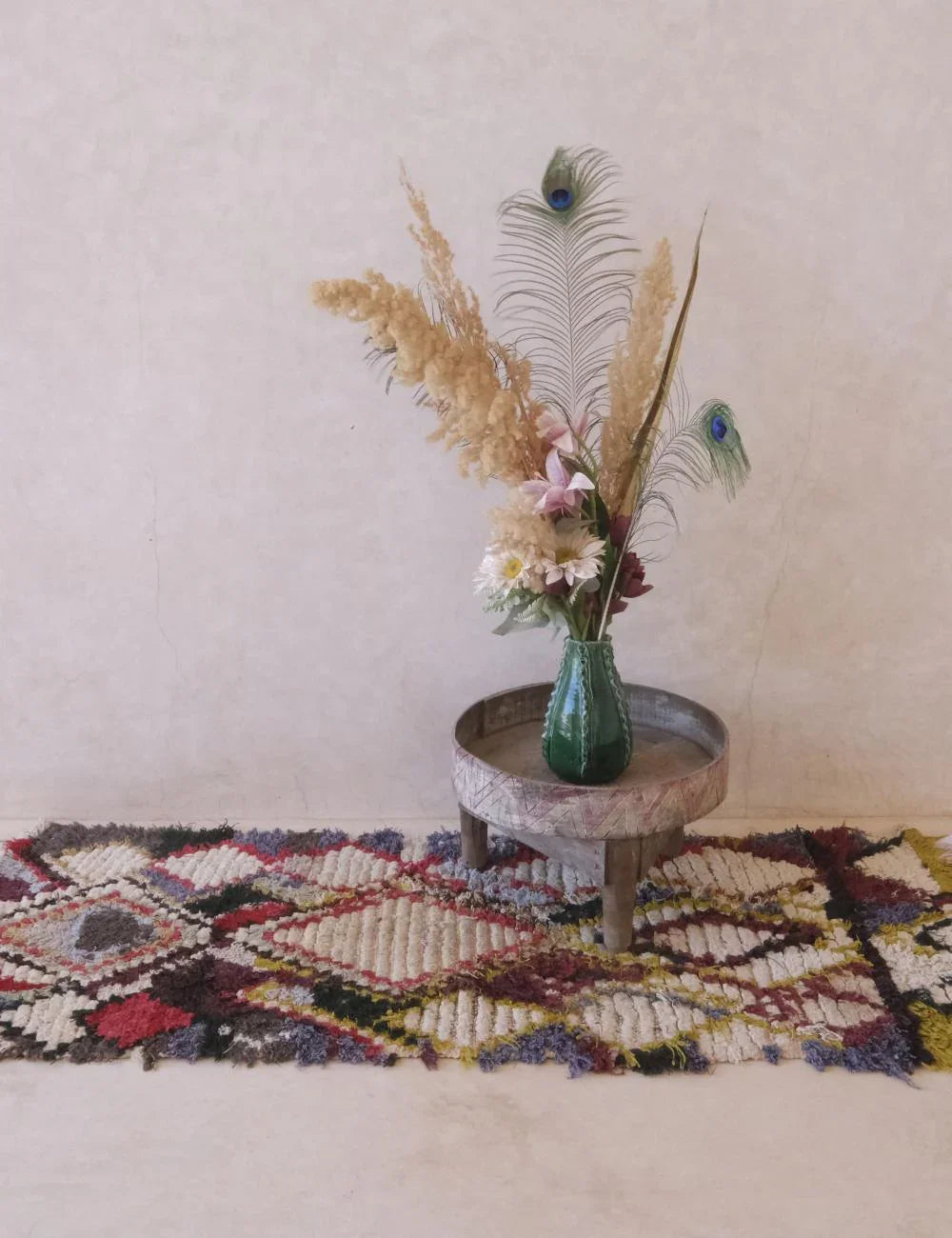 Decorative vase with flowers and peacock feathers on a small table, placed on a colorful rug against a light wall.