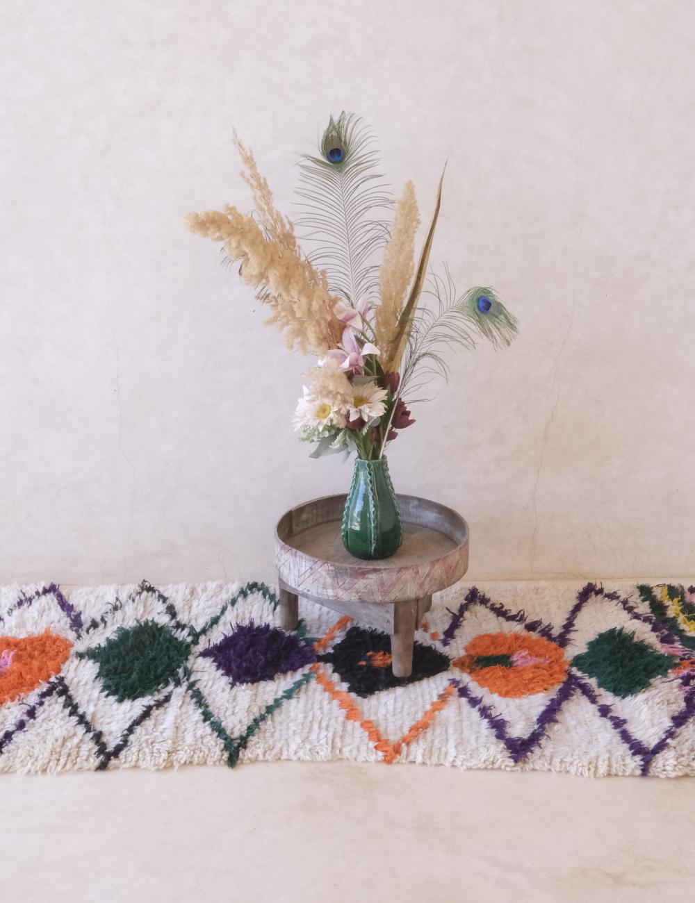 Small round wooden table with a vase of flowers and peacock feathers on a colorful rug.