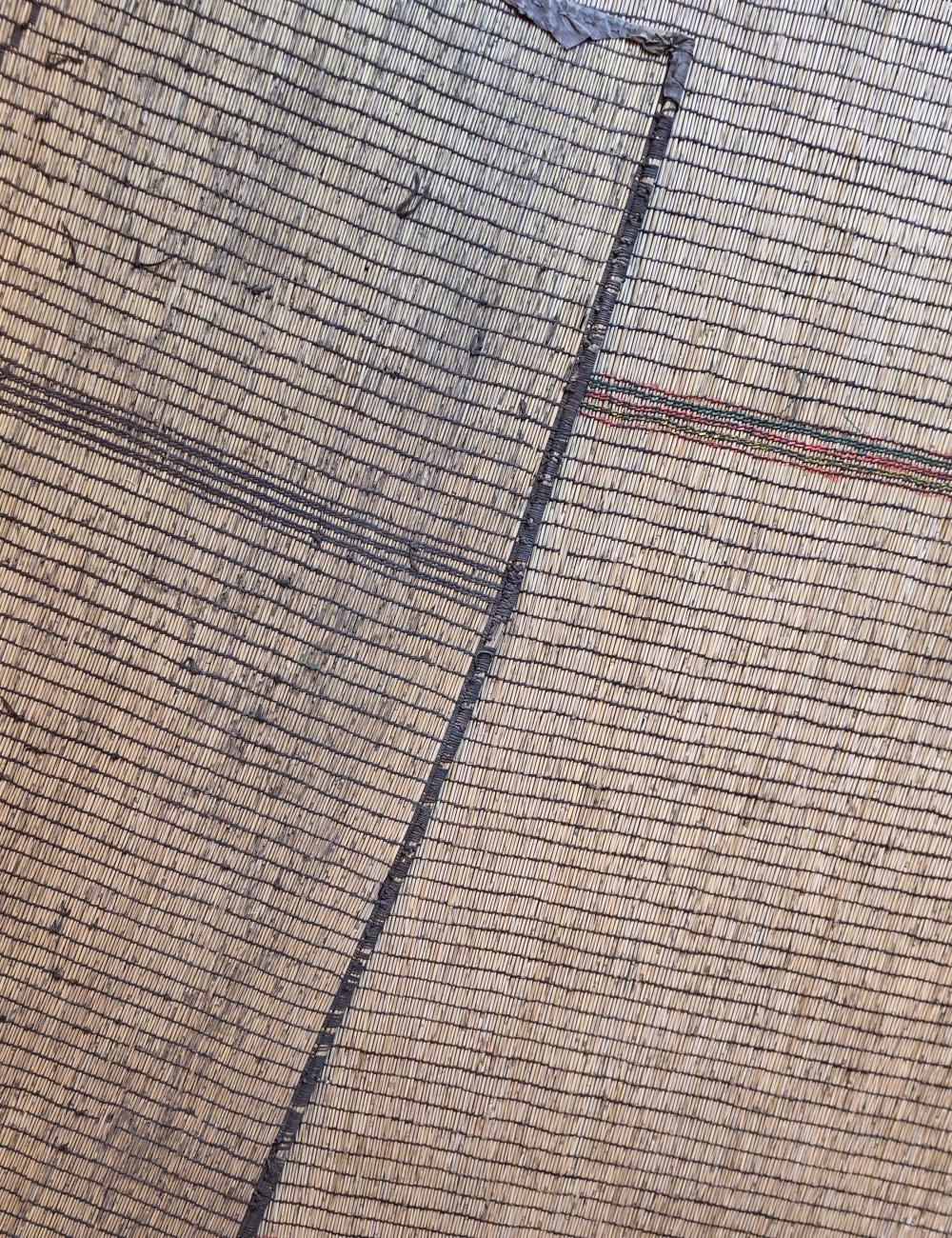 A close-up of a woven textile or rug showing a tan and brown grid pattern with dark stitched seams.