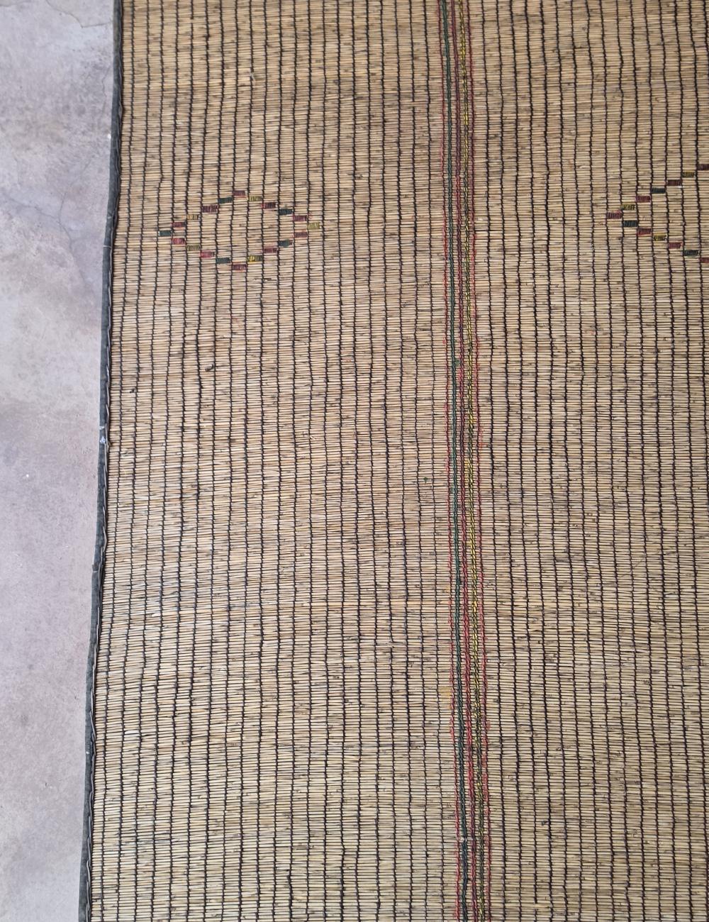 A close-up of a woven natural fiber rug or mat with vertical black stripes, a central reddish-brown stripe, and small diamond motifs near the top, shown against a gray floor background.
