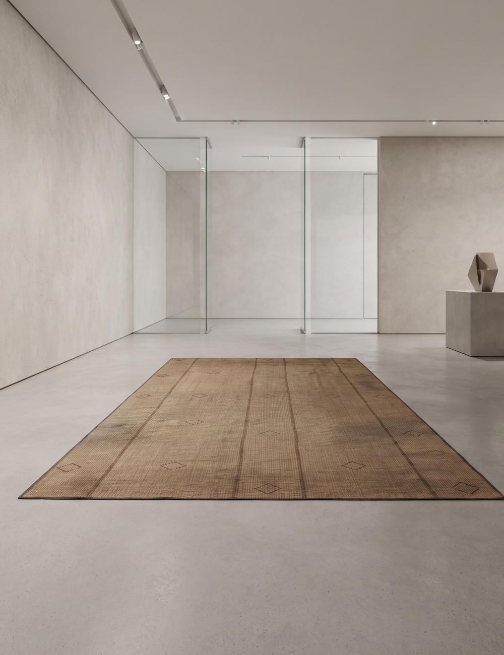 A rectangular vintage-style brown area rug with a rustic, weathered texture, displayed on a minimalist concrete floor in a modern gallery space.